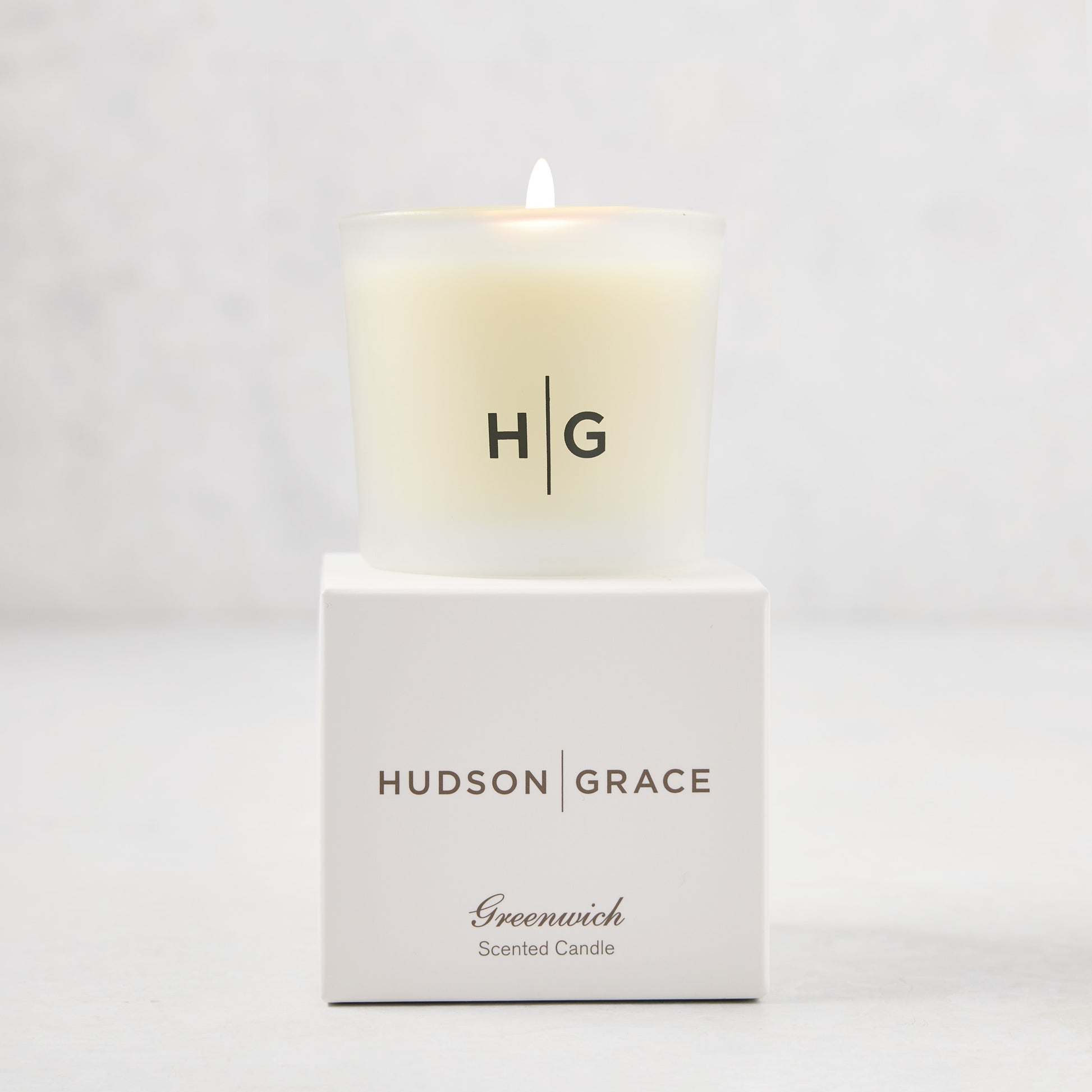 Hudson Grace Greenwich Scented Votive, a lit candle in a frosted glass vessel with "HG" logo, on its white box.