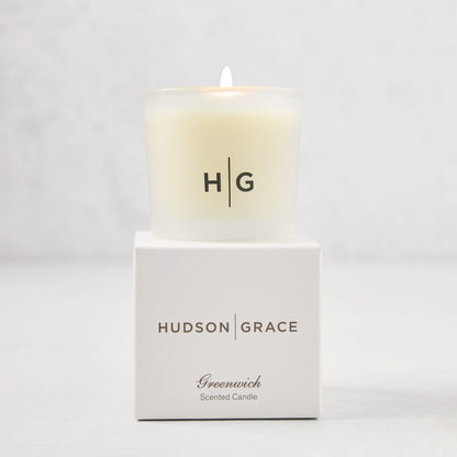 Hudson Grace Greenwich Scented Votive, a lit candle in a frosted glass vessel with "HG" logo, on its white box.