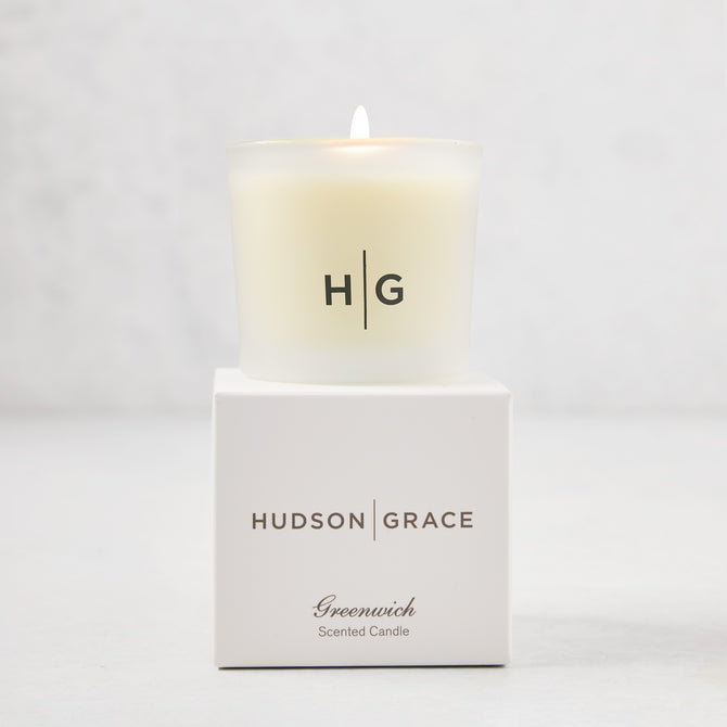 Hudson Grace Greenwich Scented Votive, a lit candle in a frosted glass vessel with "HG" logo, on its white box.