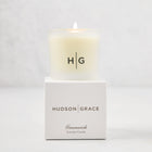 Hudson Grace Greenwich Scented Votive, a lit candle in a frosted glass vessel with "HG" logo, on its white box.