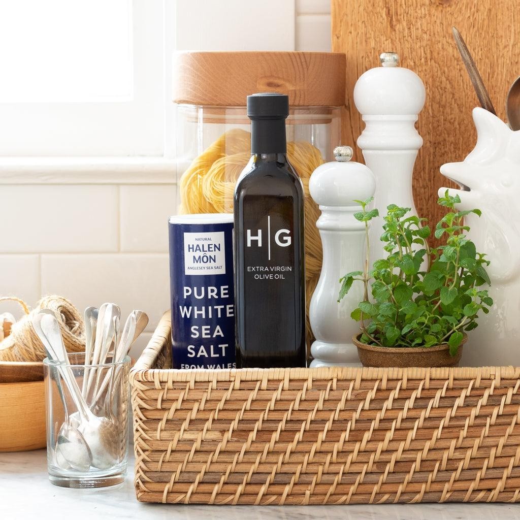 Black Hudson Grace olive oil bottle, blue sea salt container, white grinders, and herbs in a woven basket.