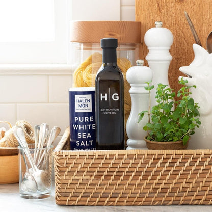 Black Hudson Grace olive oil bottle, blue sea salt container, white grinders, and herbs in a woven basket.