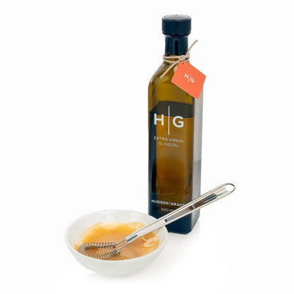 Hudson Grace Extra Virgin Olive Oil in a dark glass bottle with a small white bowl and whisk.