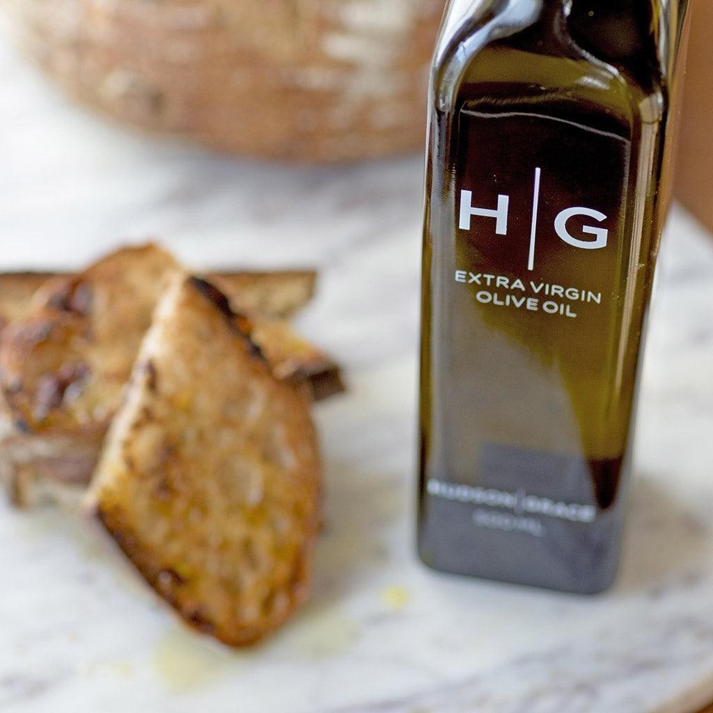 Hudson Grace Extra Virgin Olive Oil in a dark glass bottle with white HG logo, next to toasted bread slices.