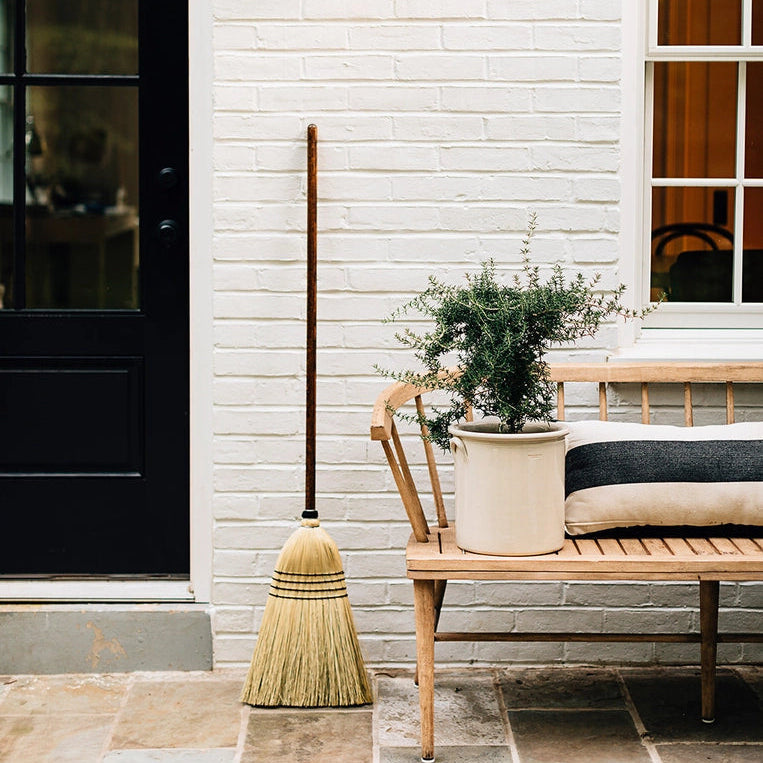 Hudson Grace handmade natural straw broom with an oak wood handle leaning against a white brick wall on a patio.