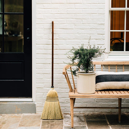 Hudson Grace handmade natural straw broom with an oak wood handle leaning against a white brick wall on a patio.