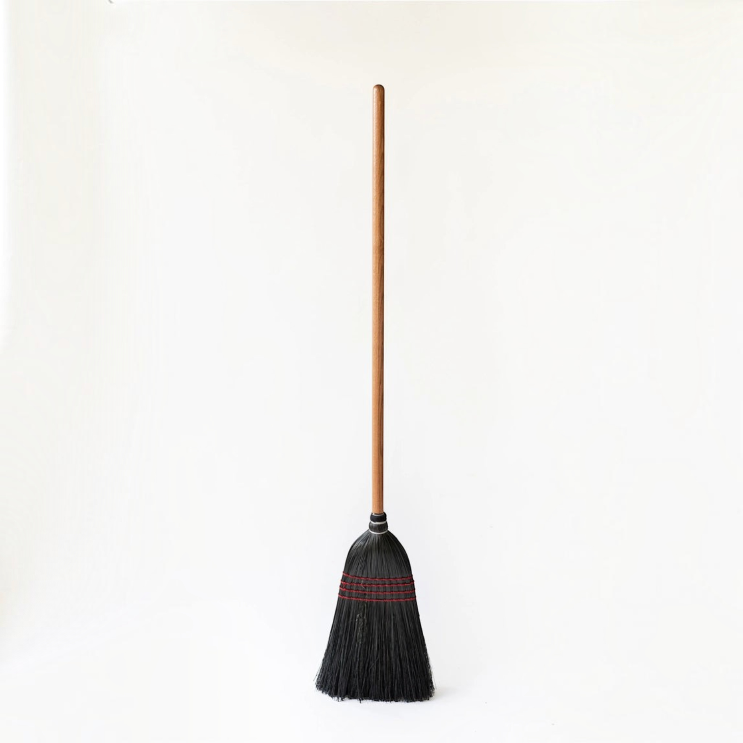 Hudson Grace handmade broom featuring a long oak wood handle and tightly bound black straw bristles with red stitching.