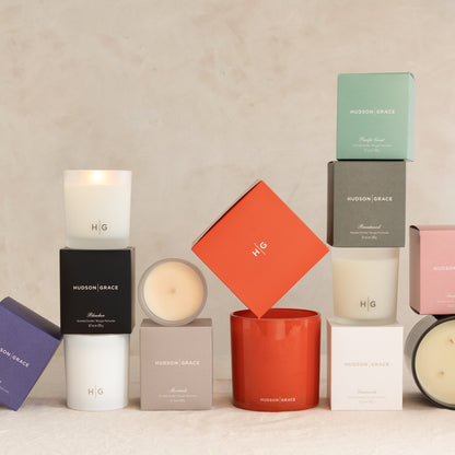 Hudson Grace scented candles in various colored boxes and glass vessels, some lit.