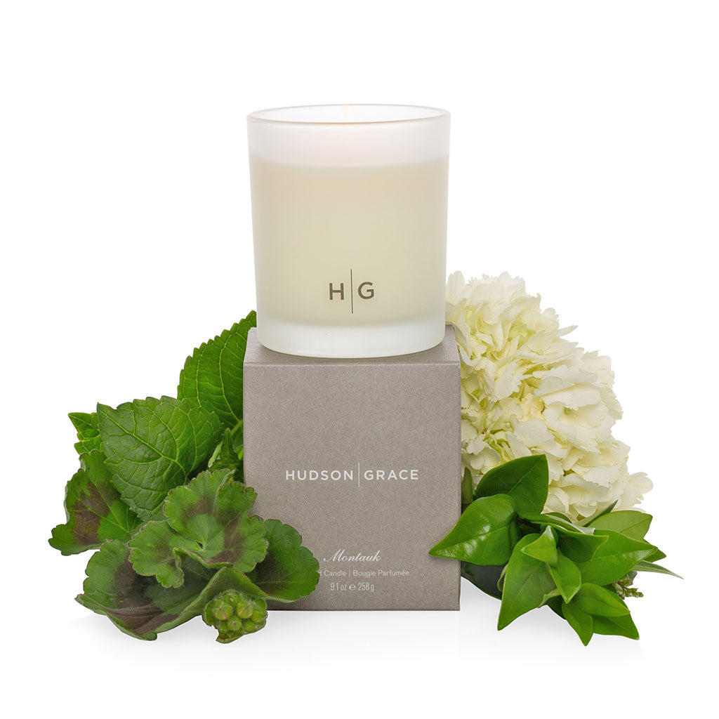 Hudson Grace Montauk scented candle in a frosted glass vessel with HG logo, on a gray box with green leaves and white flowers.