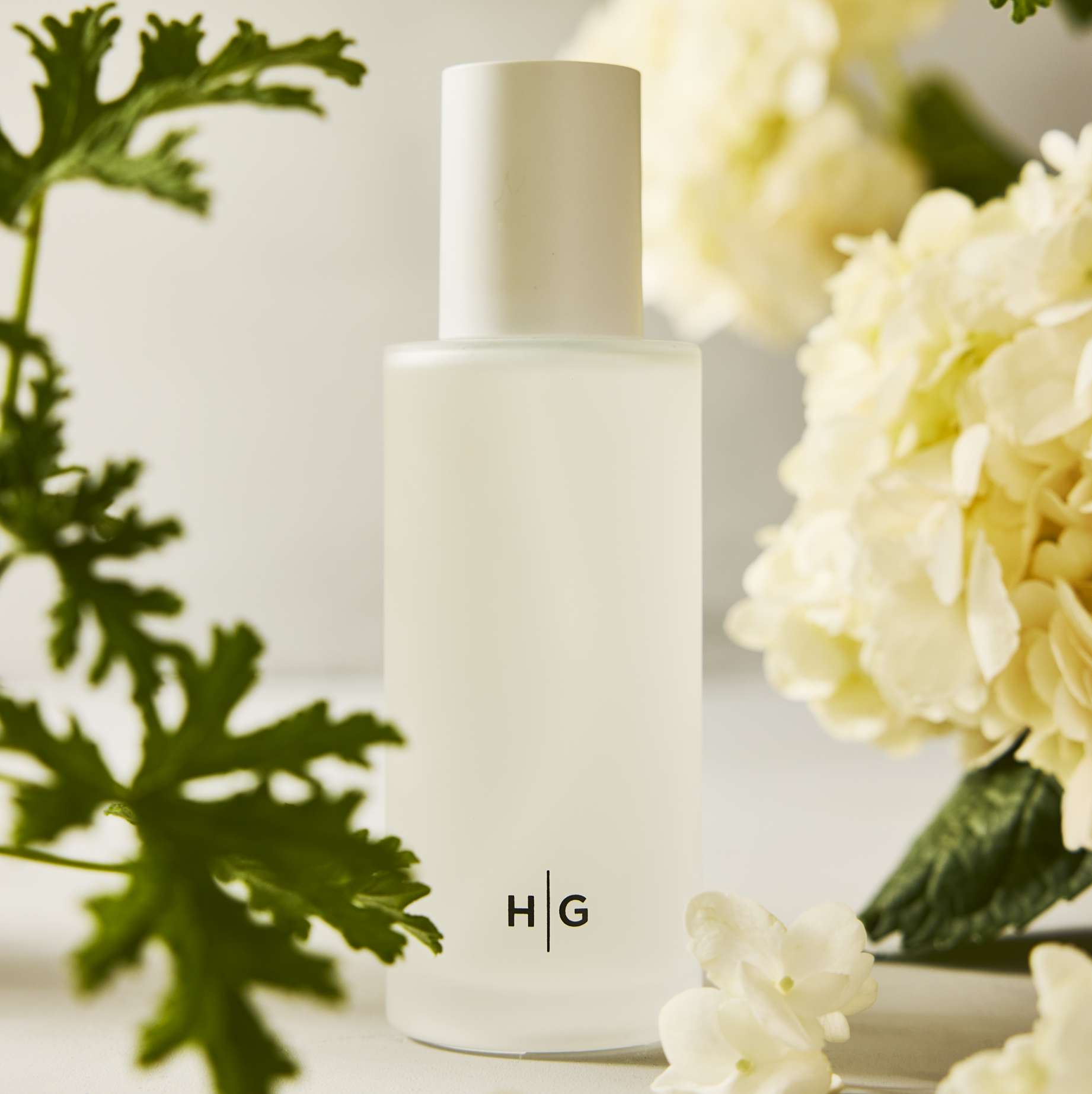 Hudson Grace Montauk Scented Reed Diffuser, frosted glass vessel with "HG" logo, surrounded by green leaves and white flowers.