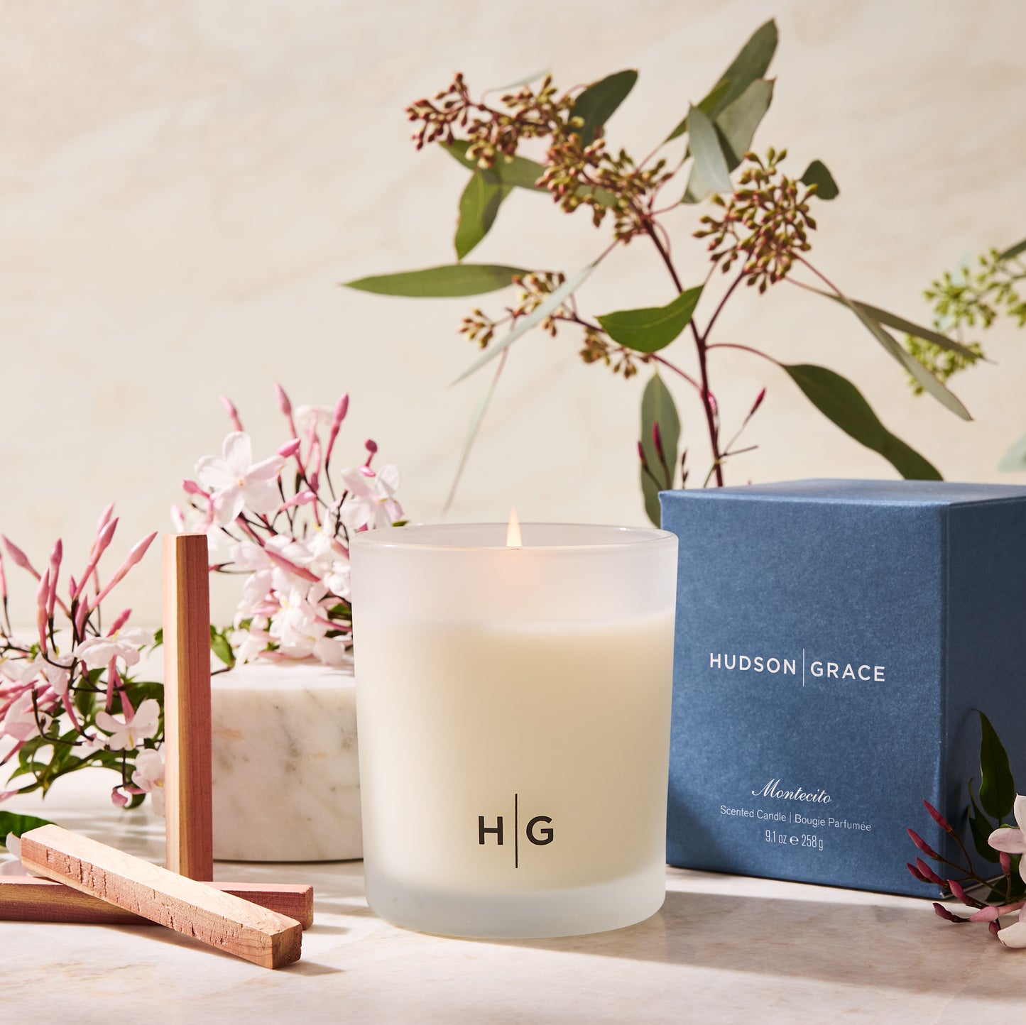 Hudson Grace Montecito Scented Candle in a frosted glass jar with a blue box, cedarwood sticks, and jasmine flowers.