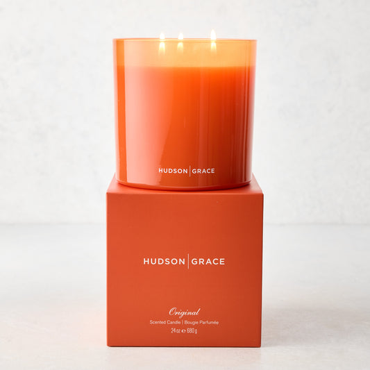 Hudson Grace Original Scented 3-Wick Candle in an orange glass vessel, lit, on its matching orange gift box.