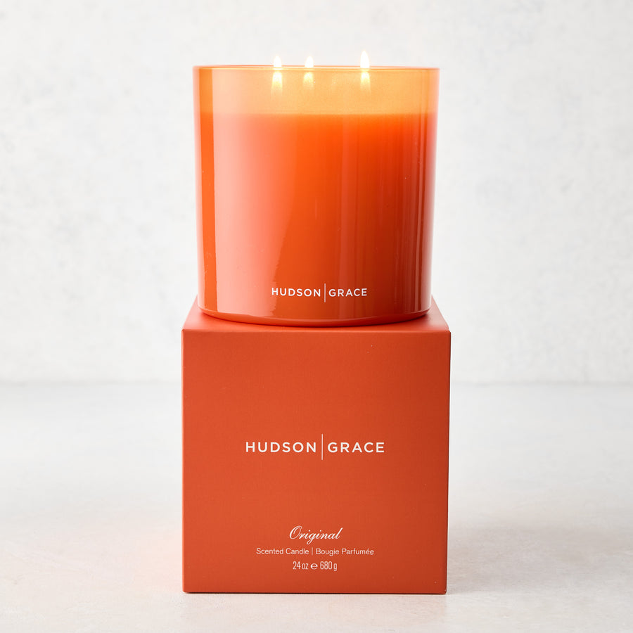 Hudson Grace Original Scented 3-Wick Candle in an orange glass vessel, lit, on its matching orange gift box.