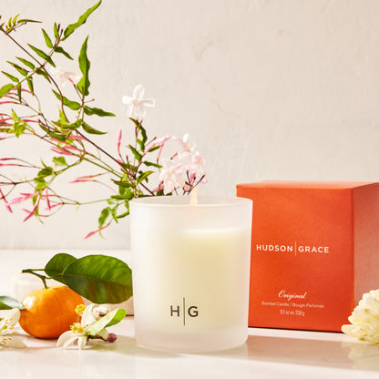 Hudson Grace Original Scented Candle, classic size, in a frosted glass vessel with a burning wick, next to an orange box.