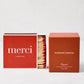 Hudson Grace Original Scented Candle in a rust-colored box next to a red "merci" matchbox with red-tipped matches.