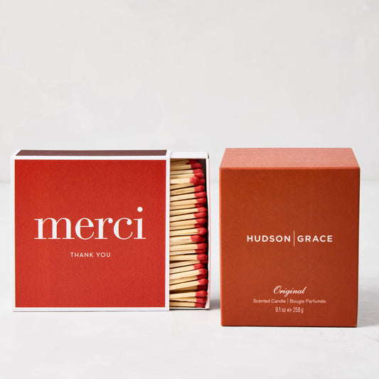 Hudson Grace Original Scented Candle in a rust-colored box next to a red "merci" matchbox with red-tipped matches.
