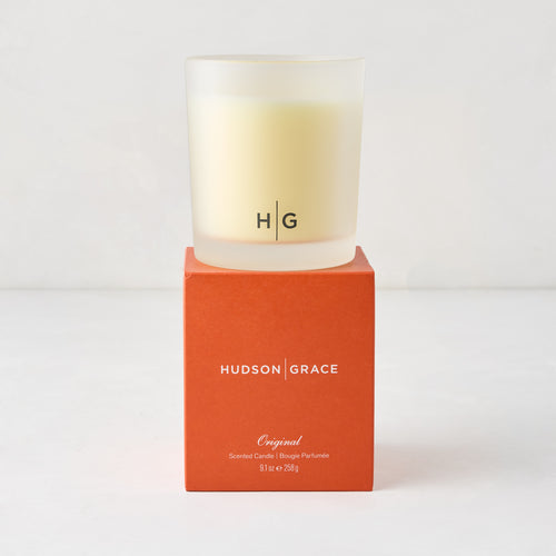 Hudson Grace Original Scented Candle in a frosted glass vessel with "HG" logo, on an orange box.