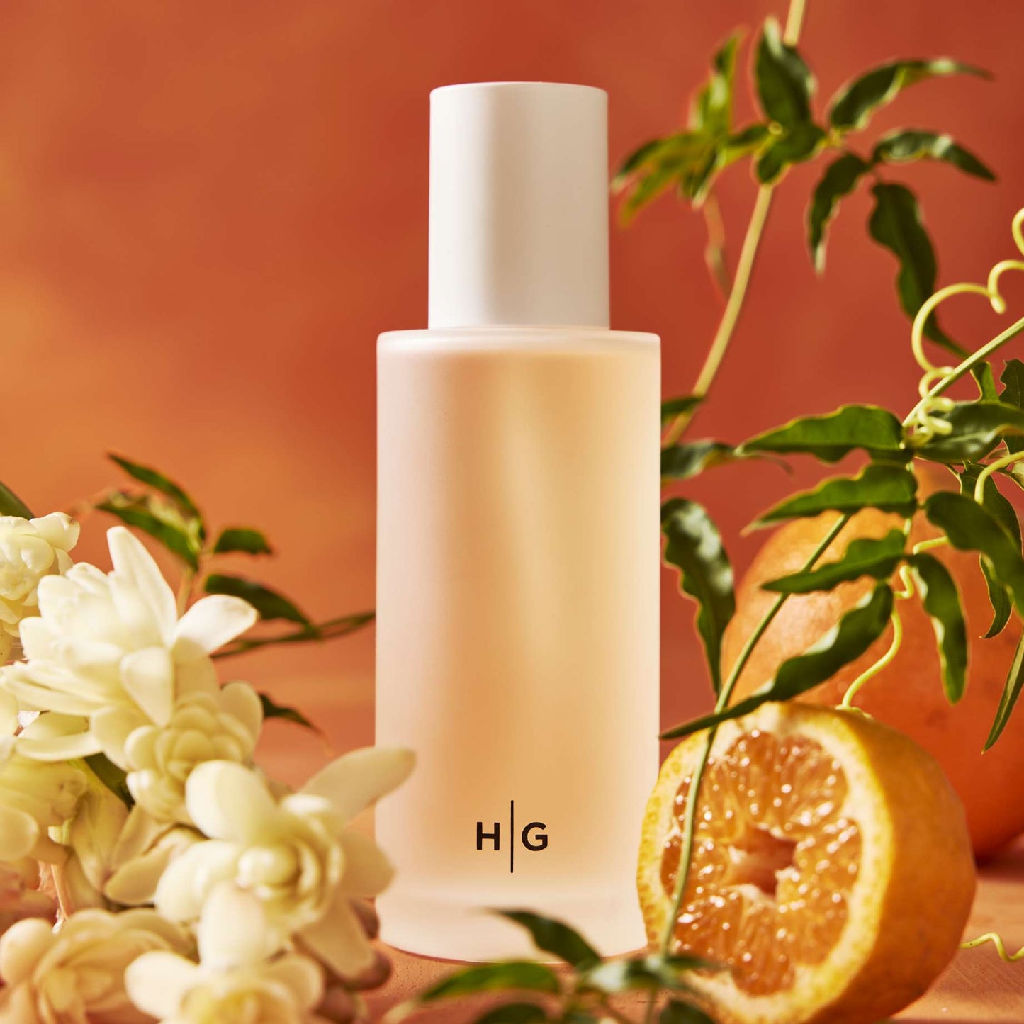 Hudson Grace Original Scented Diffuser: frosted glass bottle with "H|G" logo, white cap, surrounded by white flowers and cut oranges.