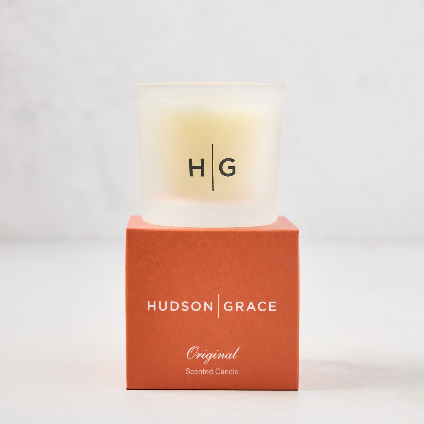 Hudson Grace Original Scented Votive Candle in a frosted glass vessel, on an orange branded box.