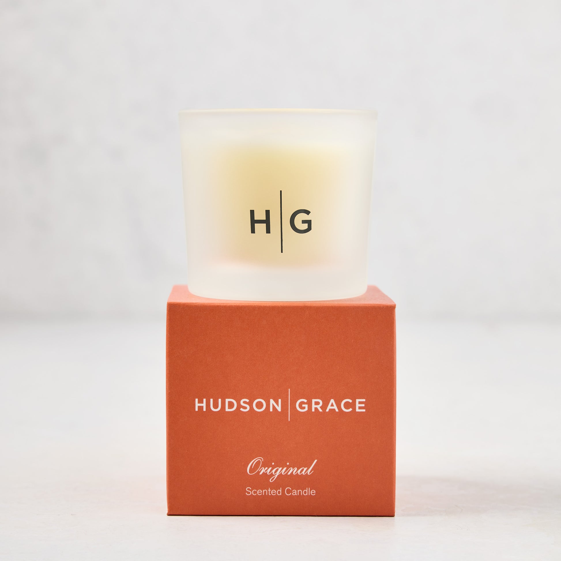 Hudson Grace Original Scented Votive Candle in a frosted glass vessel, on an orange branded box.