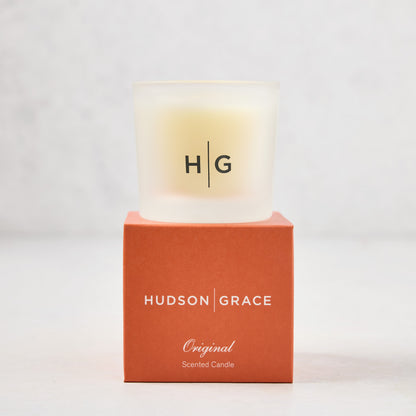 Hudson Grace Original Scented Votive Candle in a frosted glass vessel, on an orange branded box.