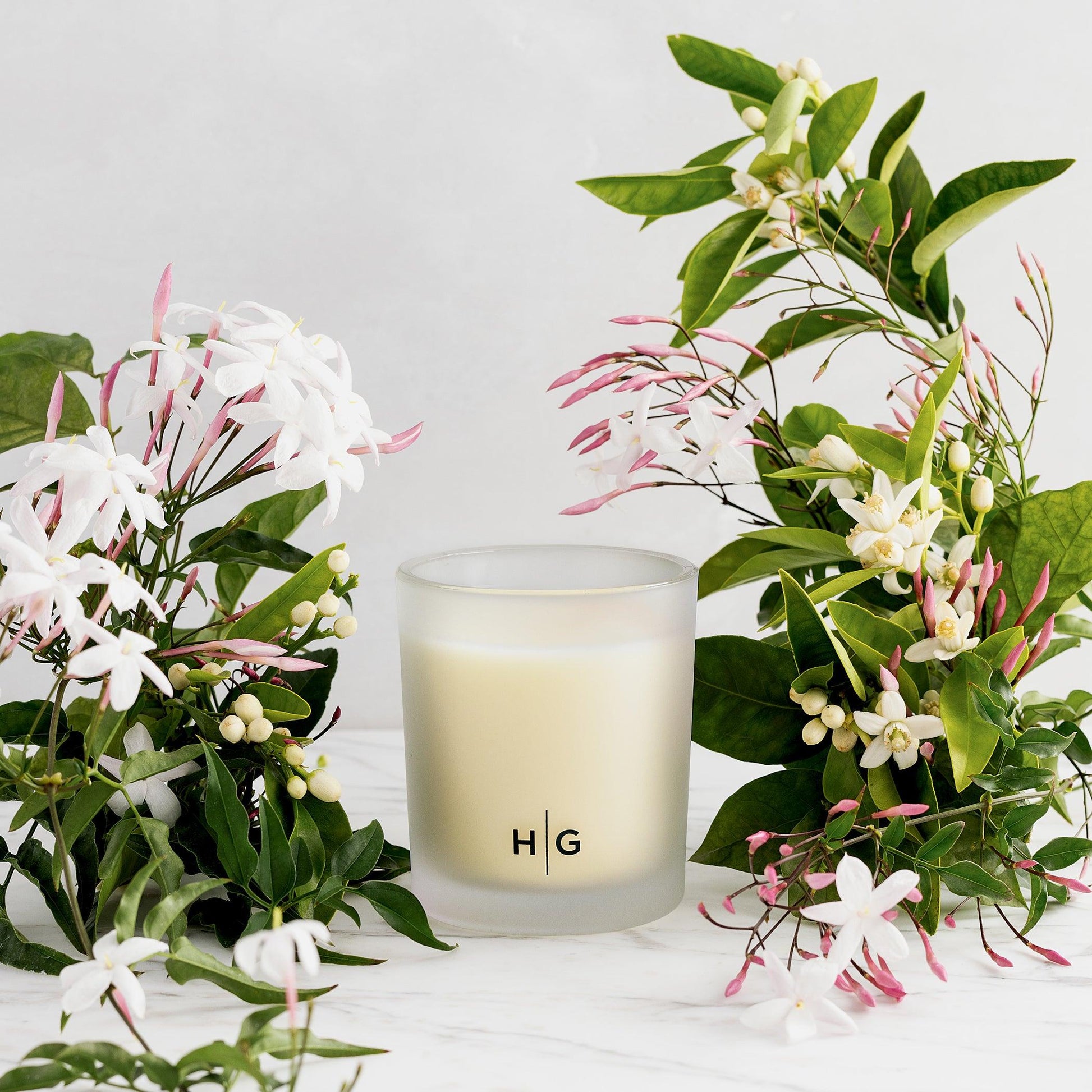Hudson Grace Original Scented Votive Candle in frosted glass with HG logo, surrounded by white and pink jasmine flowers.
