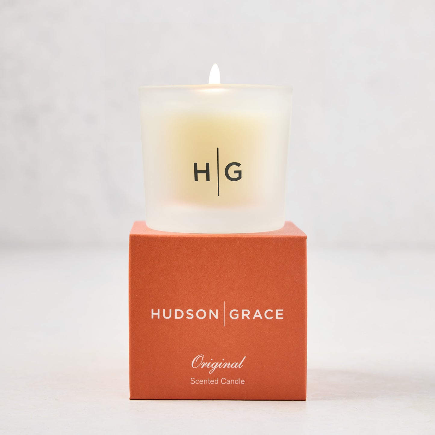 Hudson Grace Original Scented Votive Candle in a frosted glass vessel with HG logo, on an orange gift box.