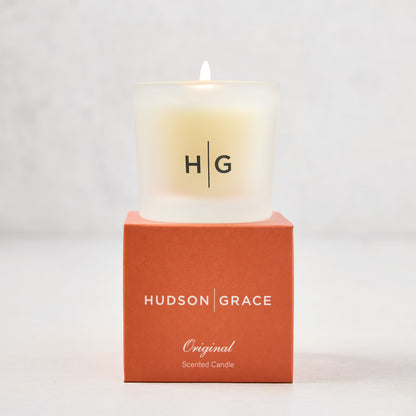 Hudson Grace Original Scented Votive Candle in a frosted glass vessel with HG logo, on an orange gift box.