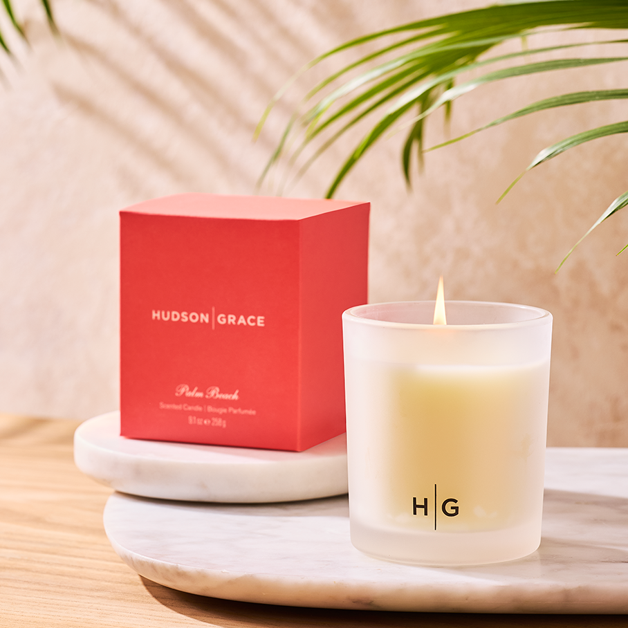 Hudson Grace Palm Beach scented candle in a frosted glass vessel with a red box, on marble with palm leaves.