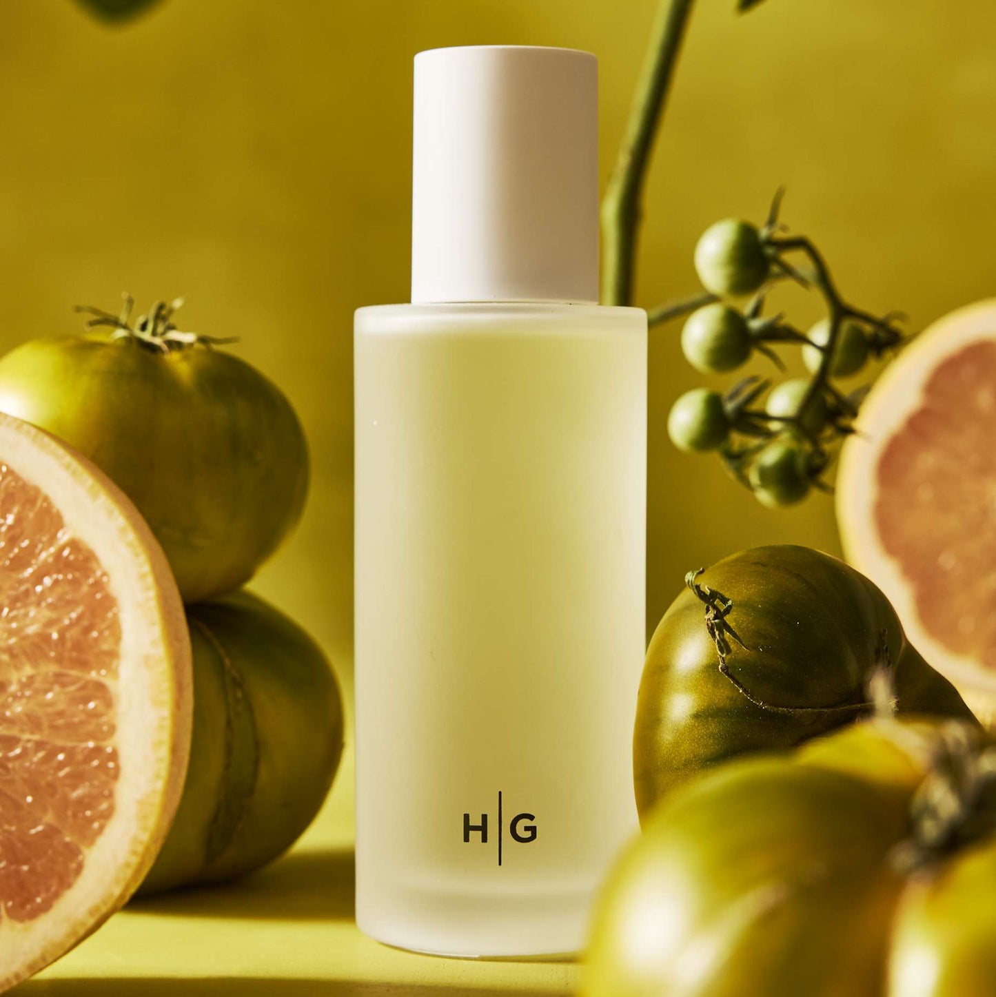 Hudson Grace San Marzano Room Spray in a minimalist frosted glass bottle with "HG" logo, surrounded by green tomatoes and grapefruit slices.