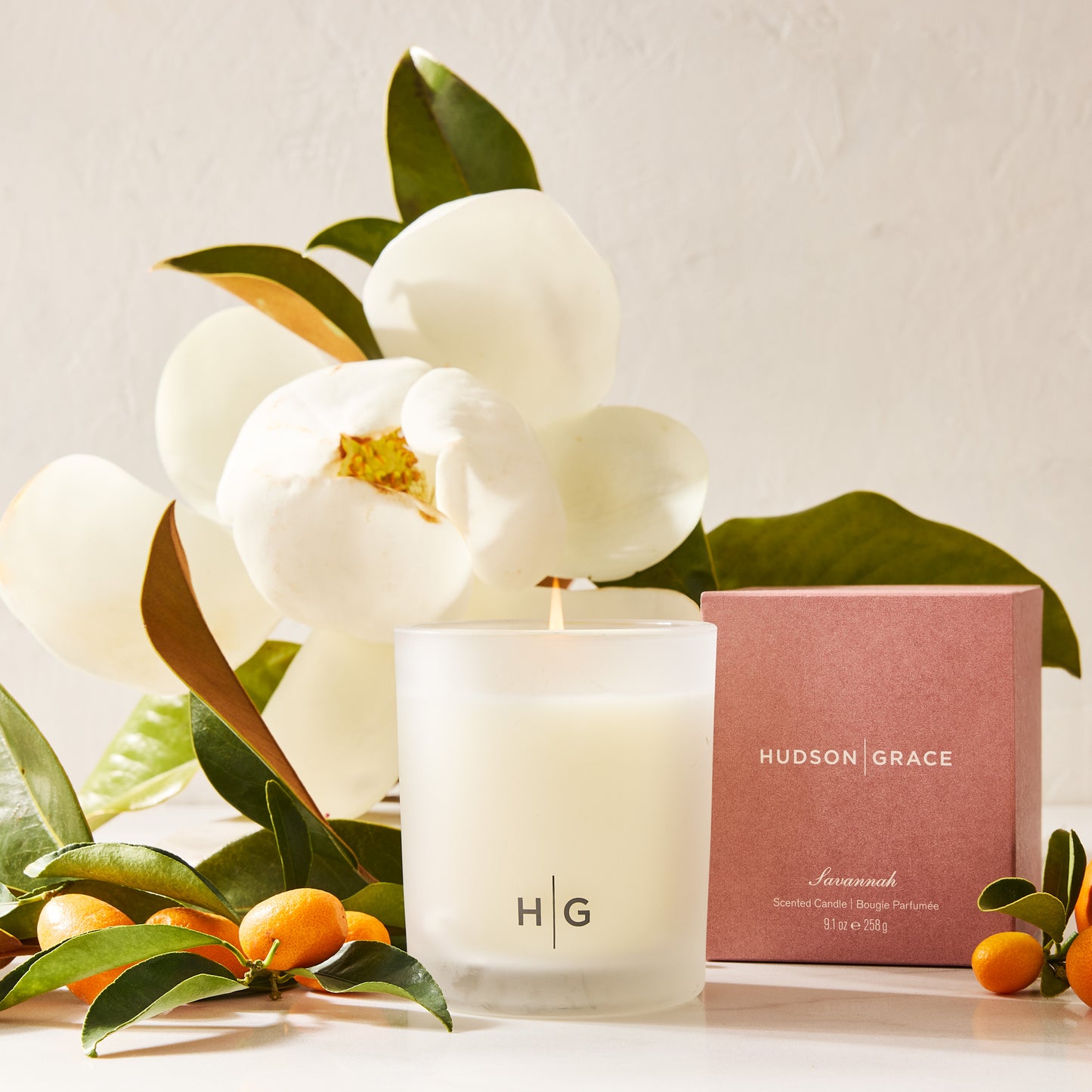 Hudson Grace Savannah Scented Candle in a frosted glass vessel with a pink box, magnolia, and kumquats.