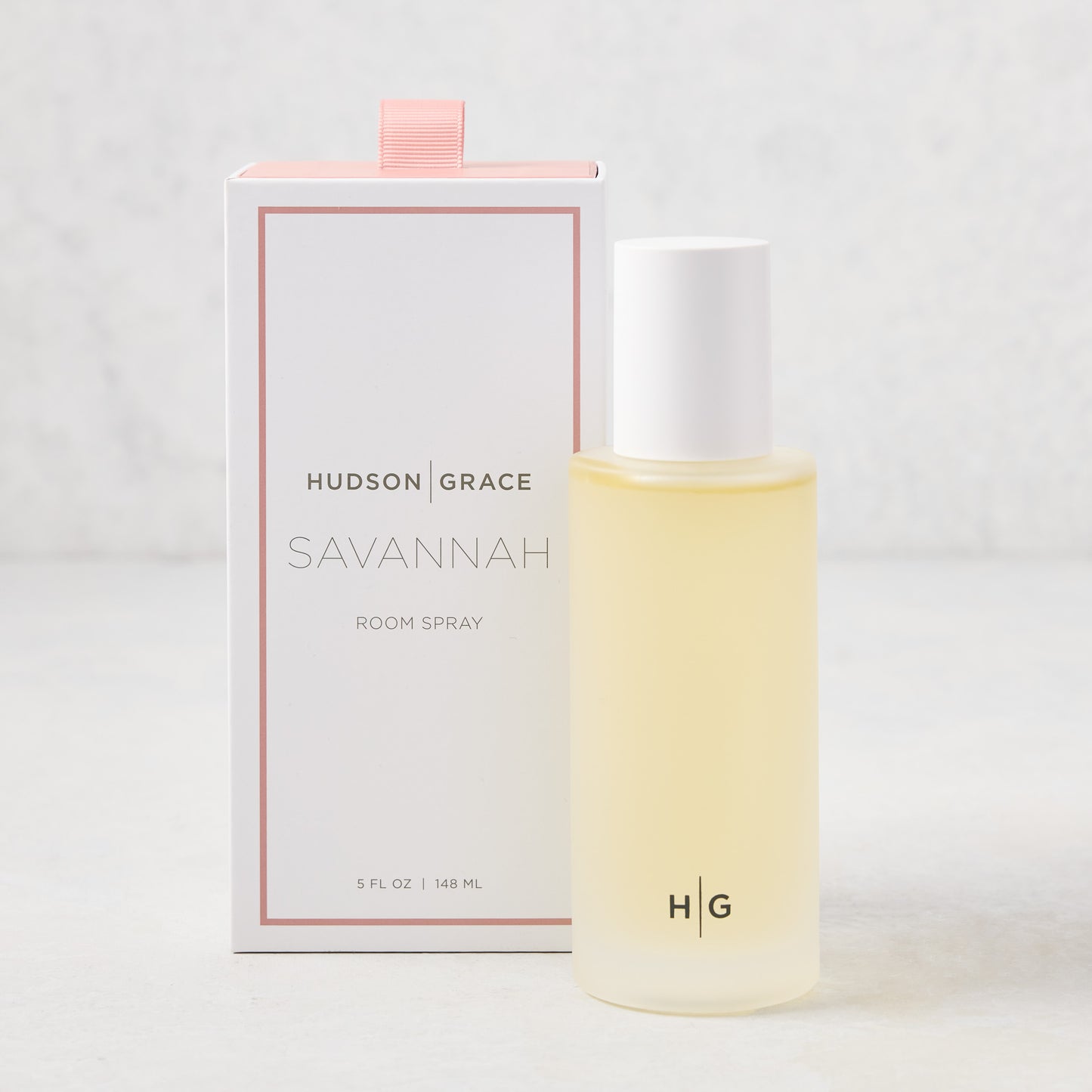 Hudson Grace Savannah Scented Room Spray: 5.08 oz clear glass bottle with white cap next to a white box with pink ribbon.