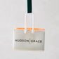 Hudson Grace shopping bag Christmas ornament, white with orange tissue, black logo, green ribbon.