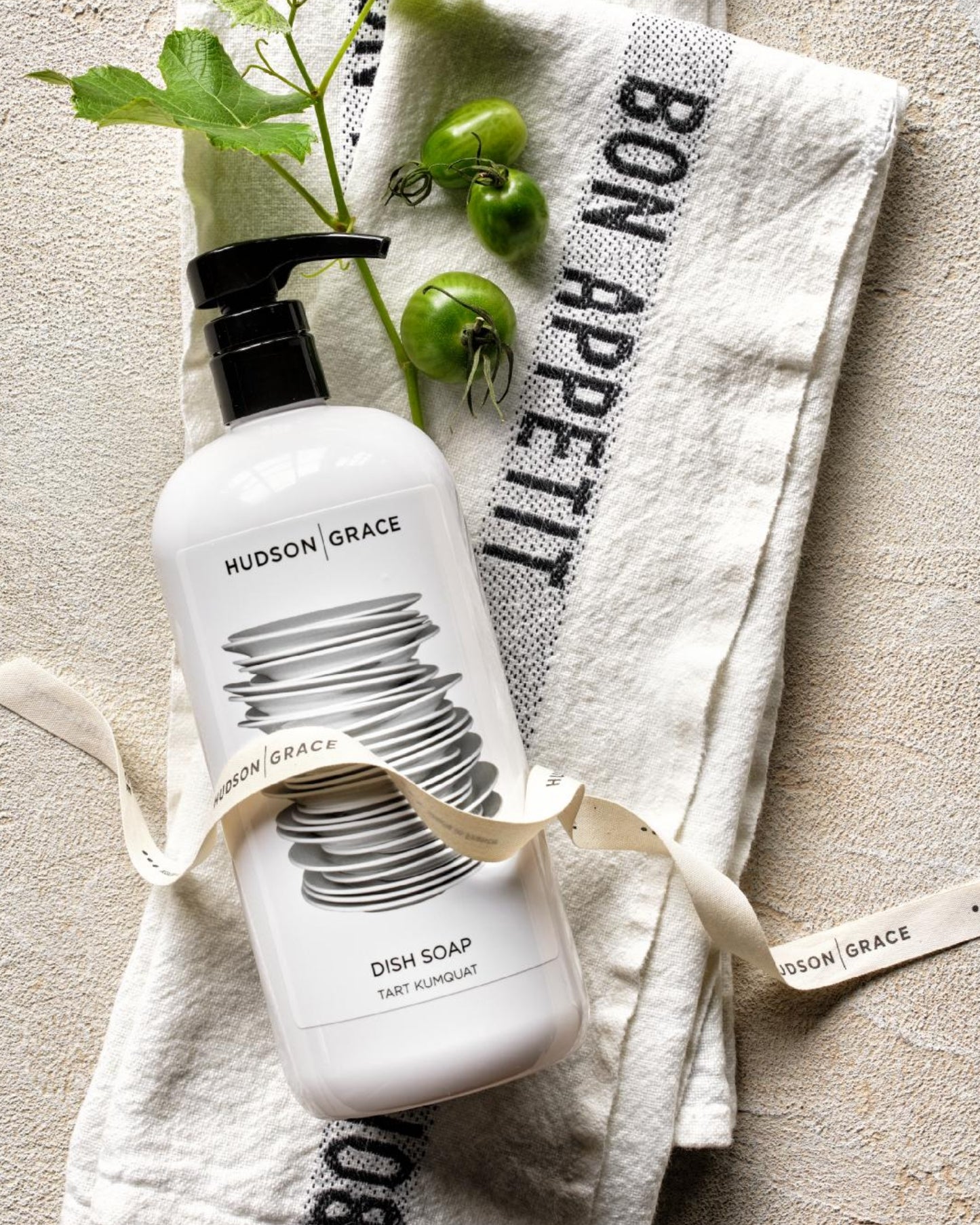 A bottle of Hudson Grace dish soap with a black pump, wrapped in a cream ribbon, styled on a textured beige towel with 'BON APPETI' printed on it, alongside green tomatoes and leaves.