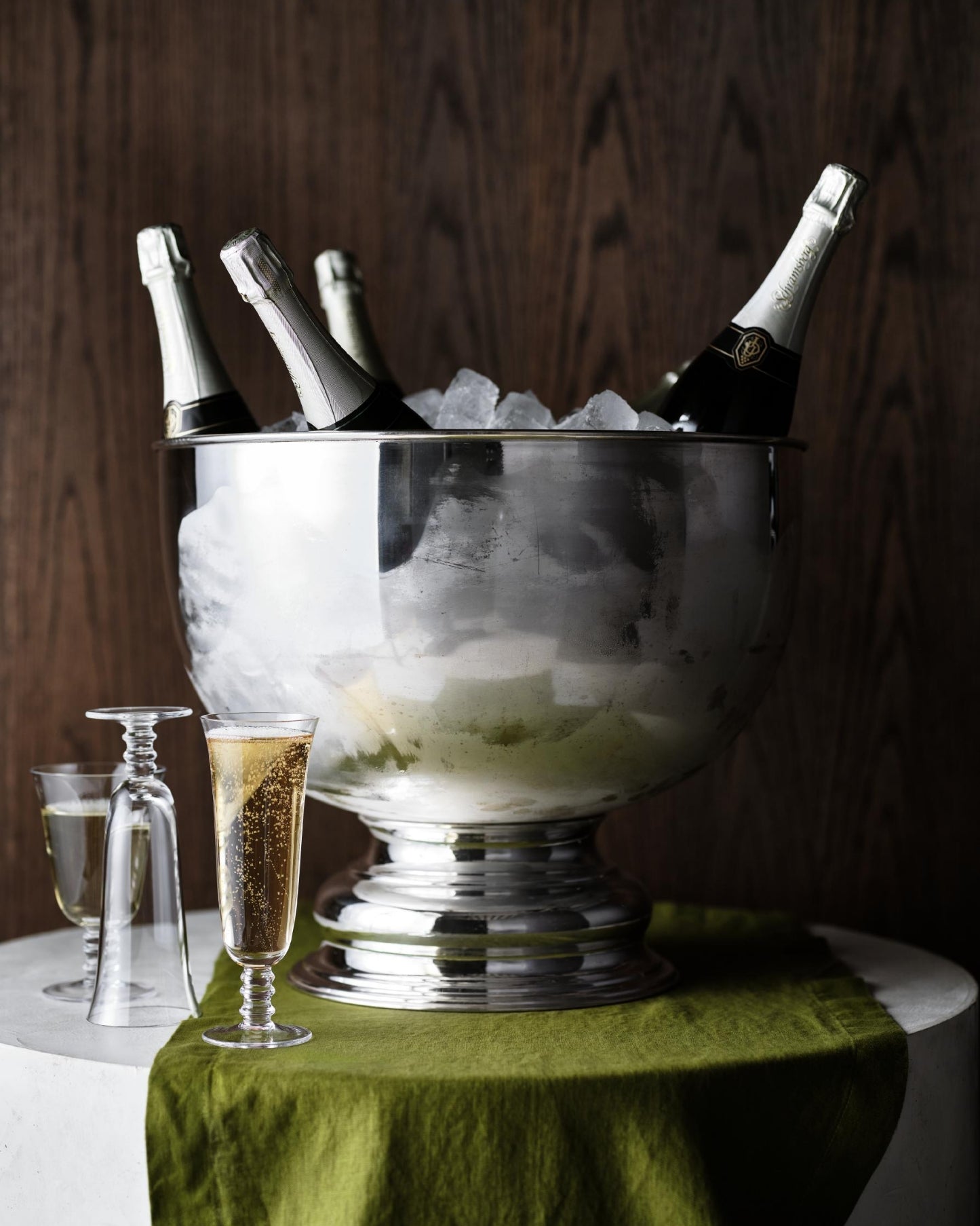 A large silver ice bucket filled with ice and champagne bottles, styled on a white table draped with an olive green cloth against a dark wood background.