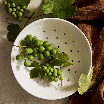 White ceramic berry bowl colander with drainage holes, holding fresh green grapes on a marble surface.