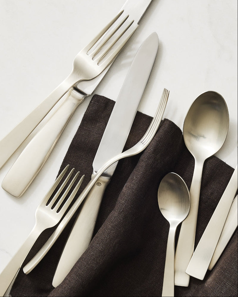 Set of silverplate flatware, including knives, forks, and spoons, on a white surface with a brown linen napkin from Hudson Grace.