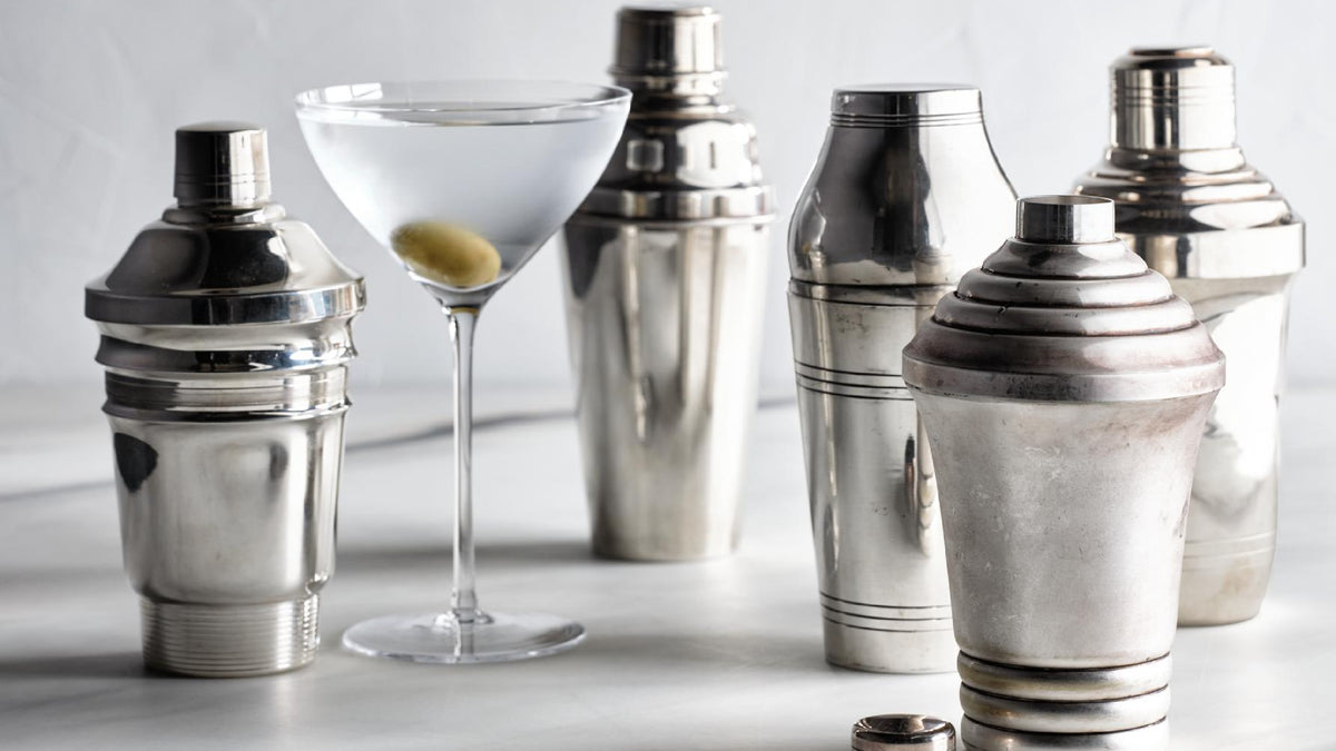 A set of polished silver metal cocktail shakers and a clear martini glass with an olive, arranged on a light surface against a soft white background.