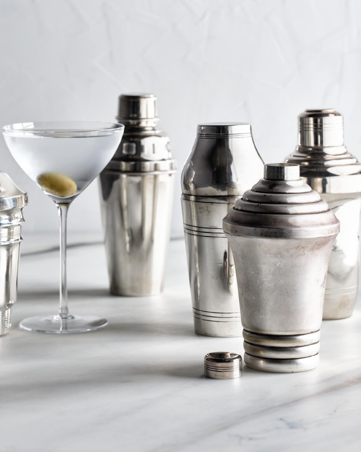 A set of polished silver metal cocktail shakers arranged on a light marble surface next to a martini glass with an olive, against a soft white textured background.