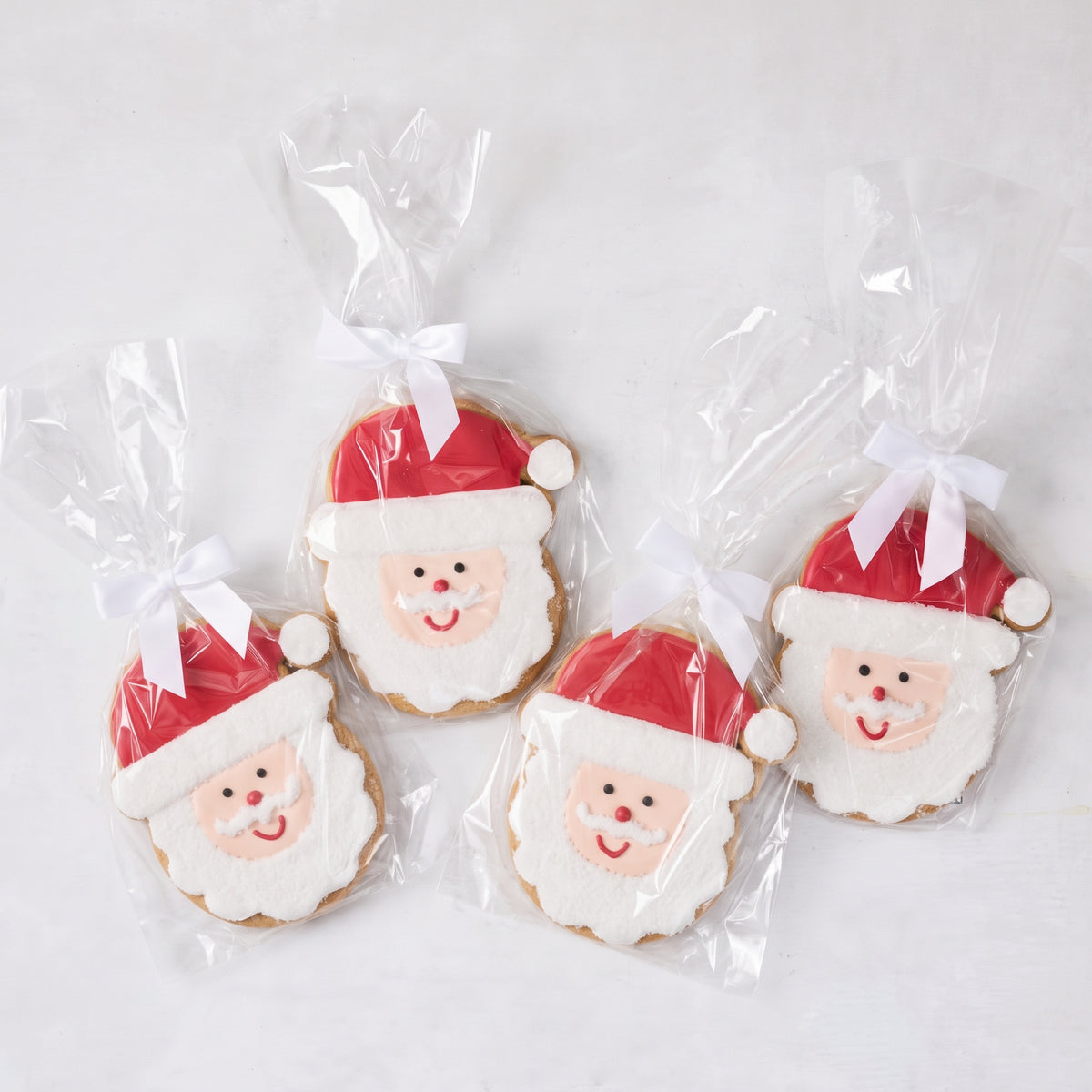 Set of four oversized sugar cookies shaped like Santa Claus, decorated with red and white icing, individually wrapped with white bows.