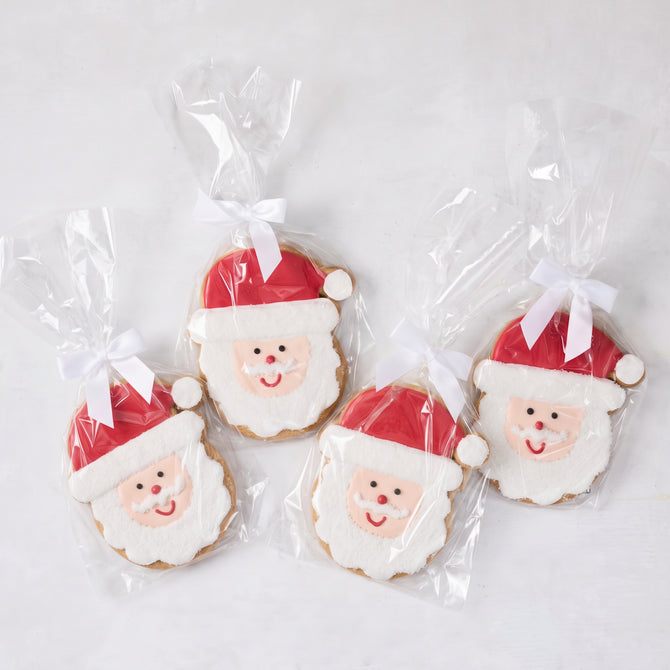Set of four oversized sugar cookies shaped like Santa Claus, decorated with red and white icing, individually wrapped with white bows.