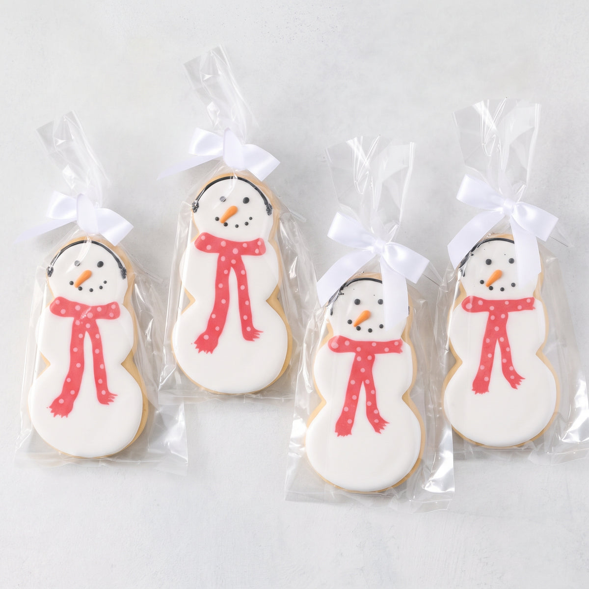 Set of four oversized snowman-shaped sugar cookies with white icing, orange carrot noses, and red polka-dot scarves in bags.