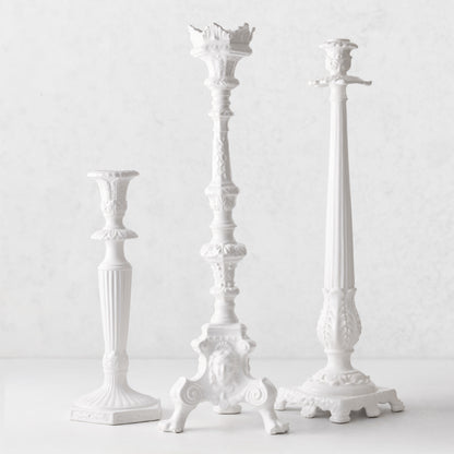 Three white Italian ceramic candlesticks with intricate antique-style detailing and footed bases on a white surface.