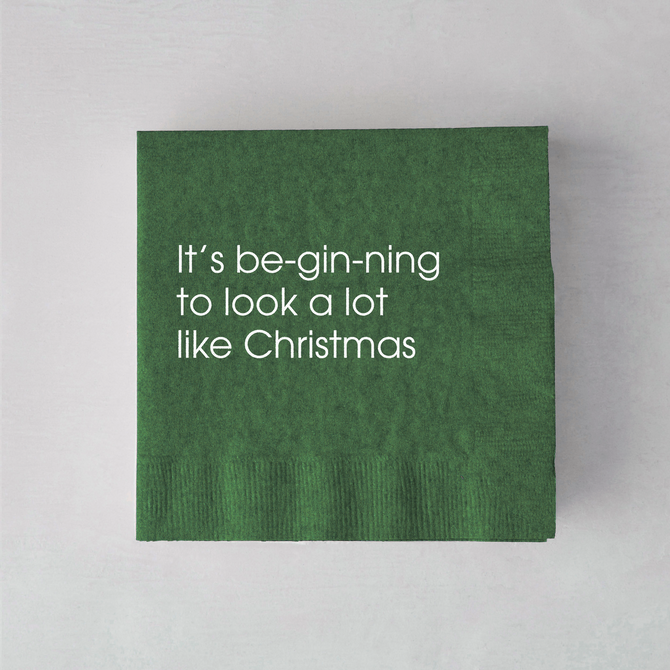 Dark green 3-ply paper cocktail napkin with white text reading It’s be-gin-ning to look a lot like Christmas.