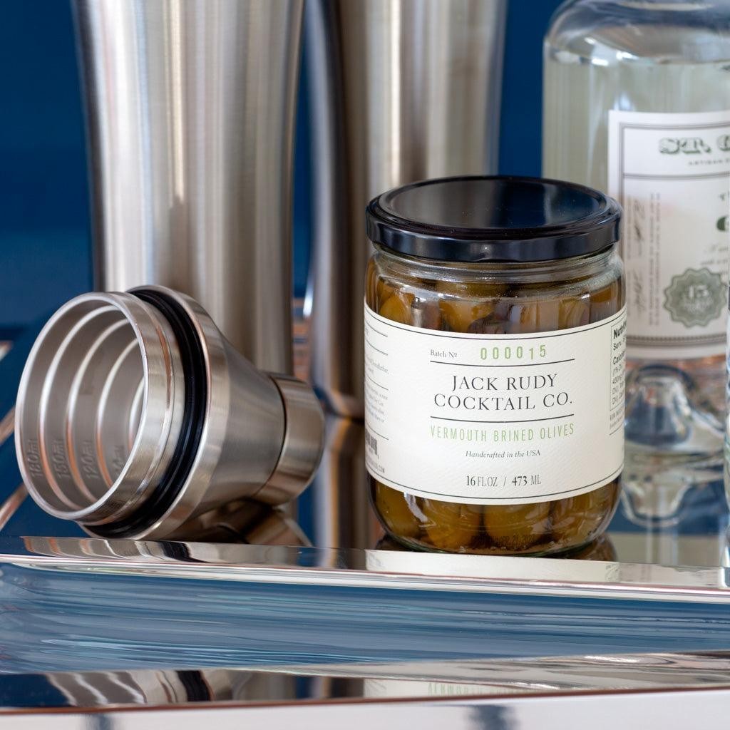 Jack Rudy Cocktail Co. Vermouth Brined Olives in a 16 fl. oz. glass jar with a black lid, on a silver tray.