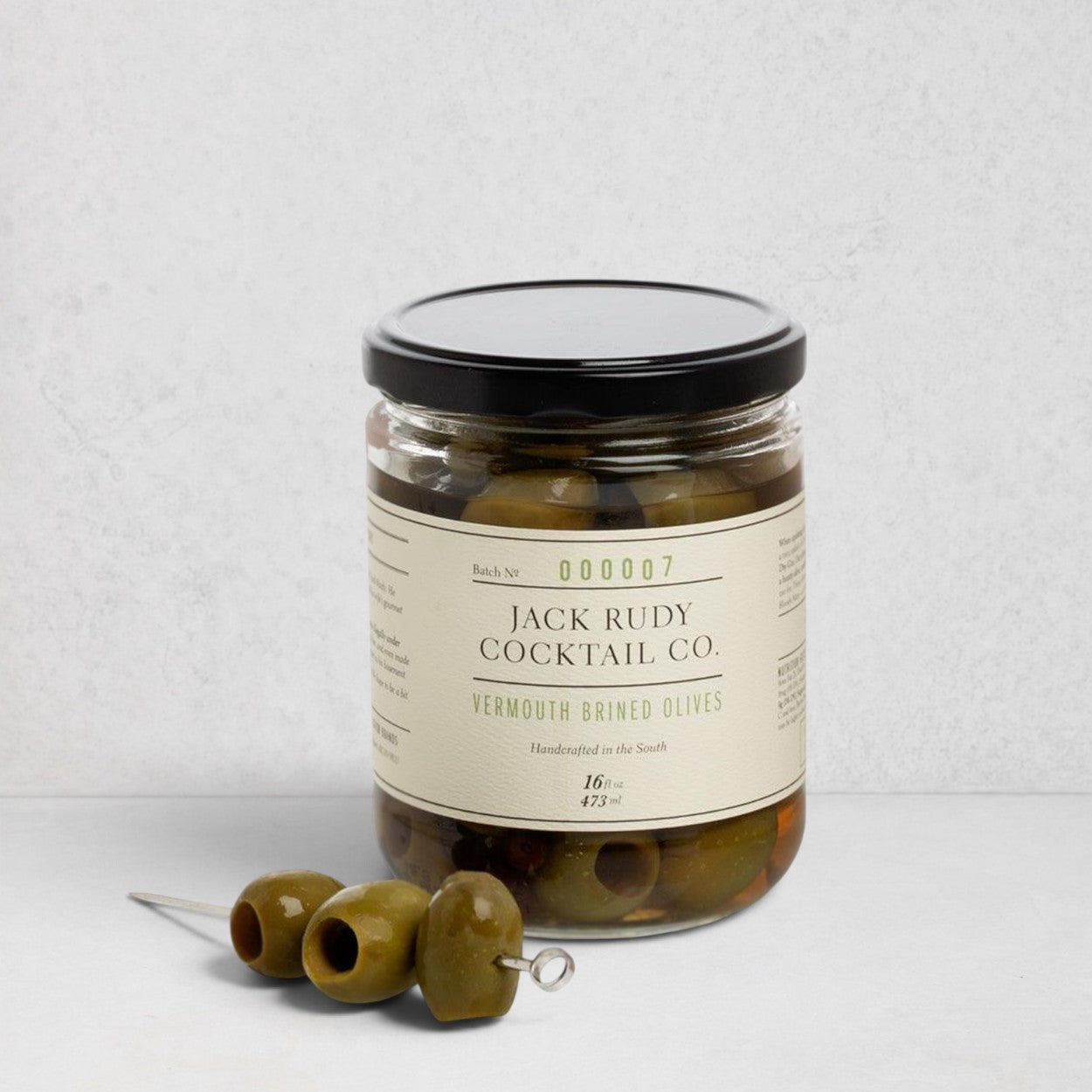 Jack Rudy Cocktail Co. Vermouth Brined Olives, 16 fl. oz. glass jar with black lid and three olives on a skewer.