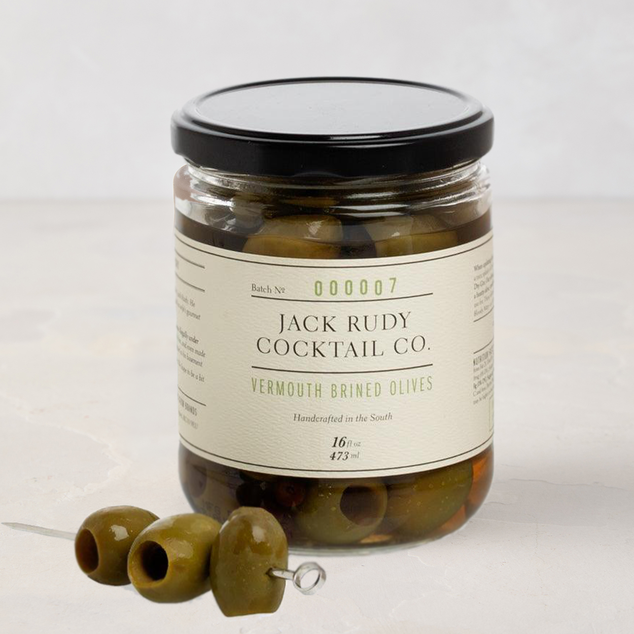 Jack Rudy Cocktail Co. Vermouth Brined Olives, 16 fl. oz. glass jar with black lid and three olives on a skewer.