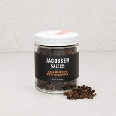 Jacobsen Salt Co. Tellicherry peppercorns in a clear glass jar with a white lid and black label on a light surface.