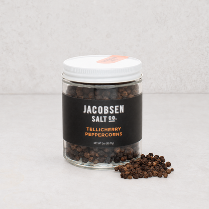 Jacobsen Salt Co. Tellicherry peppercorns in a clear glass jar with a white lid and black label on a light surface.