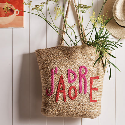 Natural jute woven tote bag with "J'ADORE" embroidered in red and pink, featuring sturdy handles.